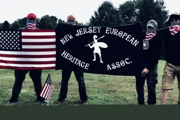 Four men wearing American flag bandanas over their faces hold a banner for the New Jersey European Heritage Association, a hate group.